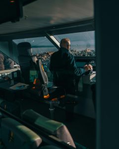 An adult male navigating a ship's control room, surrounded by seats and ocean view.